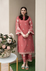 A woman wearing a pink kurta and pants stands beside a potted plant, smiling gently in a bright, natural setting.
