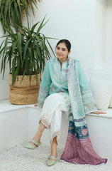 A woman dressed in a green and white outfit sits on a white wall, with a relaxed expression against a bright background.
